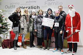 Eine Gruppe von sechs Personen hält einen großen Spendencheck über 7.350 Euro für eine gemeinnützige Organisation. Neben ihnen steht eine Person im Santa-Kostüm. Im Hintergrund sind Geschenke und ein Weihnachtsbaum sichtbar, was die festliche Atmosphäre unterstreicht.