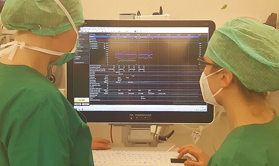 Zwei medizinisches Fachkräfte in grüner Kleidung stehen vor einem Computerbildschirm. Der Bildschirm zeigt eine Analyse mit Grafiken und Zahlen, die vermutlich medizinische Daten darstellen. Beide Personen tragen Masken und Hauben, was auf eine sterile Umgebung hinweist.