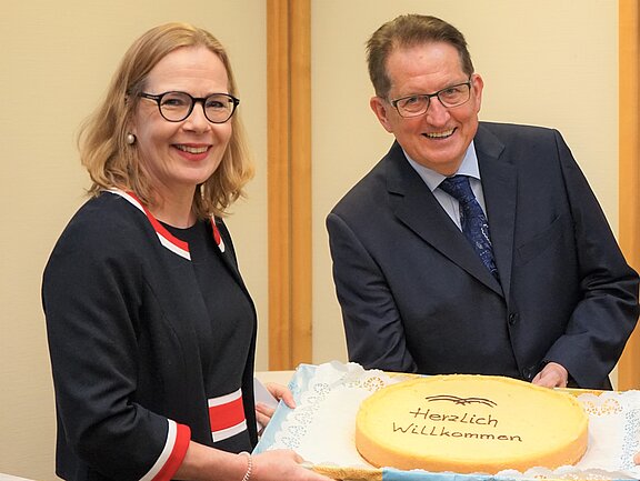 Zwei Personen lächeln freundlich in die Kamera, während sie gemeinsam eine große Torte präsentieren. Auf der Torte steht in der Mitte „Herzlich Willkommen“. Die Szene vermittelt eine herzliche Begrüßung, passend zu einem festlichen Anlass.