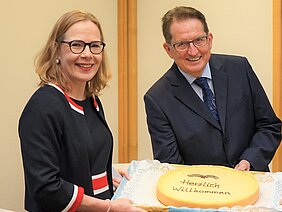 Zwei Personen lächeln freundlich in die Kamera, während sie gemeinsam eine große Torte präsentieren. Auf der Torte steht in der Mitte „Herzlich Willkommen“. Die Szene vermittelt eine herzliche Begrüßung, passend zu einem festlichen Anlass.