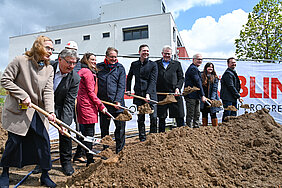 Eine Gruppe von Menschen steht an einer Baugrube und hält Schaufeln in der Hand. Sie sind bereit, den ersten Spatenstich zu setzen. Im Hintergrund ist ein Banner mit dem Wort "BAUBEGINN" sichtbar. Es ist ein sonniger Tag mit etwas bewölktem Himmel.