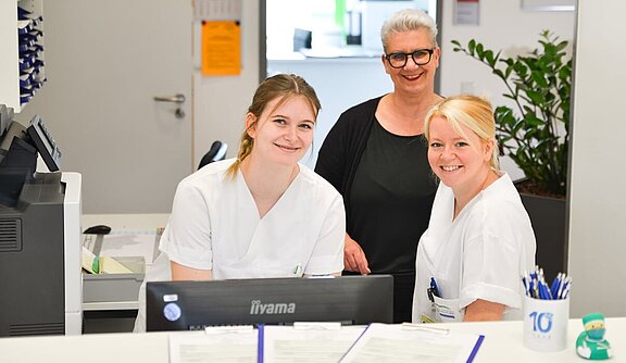 Drei Mitarbeiterinnen stehen lächelnd an einem Empfangstresen. Sie tragen weiße Kittel und sind umgeben von Schreibtischmaterialien. Im Hintergrund sind Pflanzen und ein PC sichtbar. Die Atmosphäre ist freundlich und einladend.