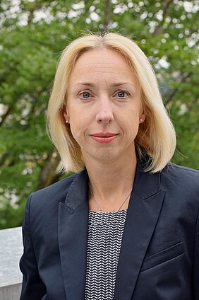 Porträt einer blonden Frau mit kurzen Haaren, die einen blauen Blazer und ein gestreiftes Oberteil trägt. Sie steht im Freien vor einer grünen Umgebung und schaut direkt in die Kamera.