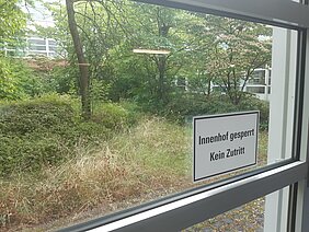 Ein Schild ist an einem Fenster angebracht und informiert: "Innenhof gesperrt. Kein Zutritt." Im Hintergrund sind grüne Bäume und Sträucher zu sehen, die eine natürliche, aber unordentliche Umgebung darstellen.