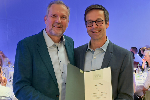 Zwei Männer stehen nebeneinander und lächeln. Der links trägt einen blauen Blazer und hat einen Bart, der rechts einen grauen Anzug und Brille. Sie halten ein Zertifikat in der Hand, das einen Preis oder eine Auszeichnung repräsentiert. Im Hintergrund sind Menschen an einem Tisch sichtbar.