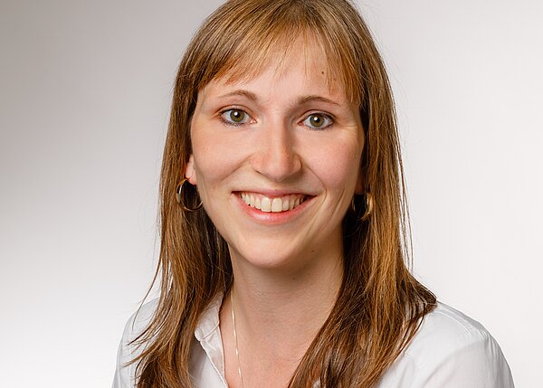 Porträt einer lächelnden Frau mit langen, braunen Haaren, die einen weißen Hemdkragen trägt. Der Hintergrund ist hell und neutral. Ihr Gesichtsausdruck ist freundlich und einladend.