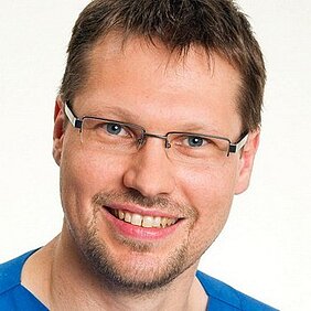 Ein lächelnder Mann mit Brille trägt ein blaues Oberteil. Sein Gesicht zeigt ein freundliches und offenes Ausdruck. Der Hintergrund ist neutral und hell gestaltet.