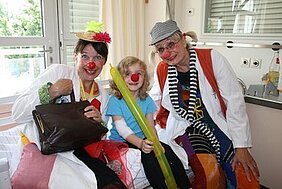 Zwei Clowns in weißen Kostümen und mit roten Nasen posieren mit einem Kind, das einen grünen Ballon in der Hand hält. Alle drei lächeln und scheinen eine fröhliche Stimmung zu haben. Im Hintergrund sind helle Wände und Fenster sichtbar.