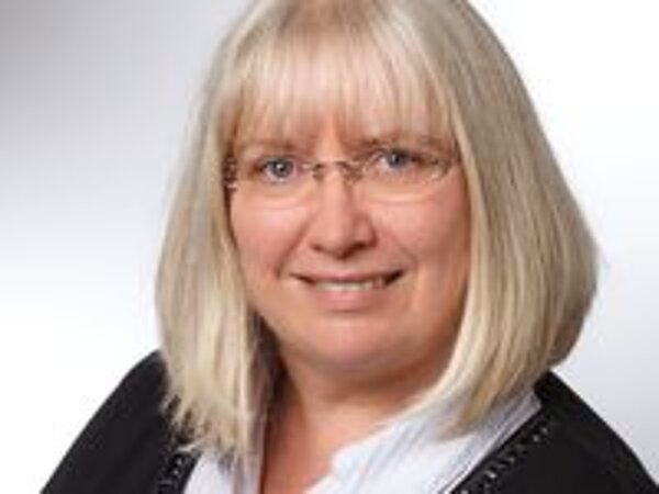 Porträt einer Frau mit blonden, schulterlangen Haaren und Brille. Sie trägt eine schwarze Jacke und lächelt freundlich in die Kamera. Der Hintergrund ist hell und verschommen.