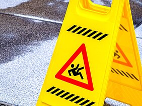 Ein gelbes Warnschild steht auf einem nassen Boden. Es zeigt ein Symbol für Rutschgefahr und warnt Passanten, vorsichtig zu sein. Der Boden ist uneben und weist eine klare Feuchtigkeit auf, was das Risiko von Stürzen erhöht.
