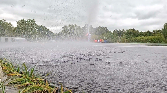 Ein Wasserstrahl spritzt aus dem Boden, während sich der Regen auf der asphaltierenden Fläche verteilt. Im Hintergrund sind grüne Bäume und verschwommene Figuren zu erkennen, die sich in einer leicht nebligen Umgebung bewegen. Der Himmel ist bewölkt.