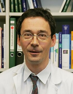 Ein Mann trägt einen weißen Laborkittel und eine Brille. Er steht vor einem Bücherregal mit verschiedenen Ordnern. Sein Gesichtsausdruck ist neutral und freundlich.