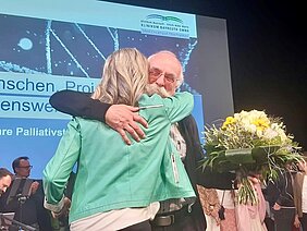 Eine Frau in einer grünen Jacke umarmt einen älteren Mann mit Brille und Bart, der einen bunten Blumenstrauß in der Hand hält. Im Hintergrund sind weitere Personen und ein Banner sichtbar, das Informationen über ein Projekt zu Palliativversorgung zeigt.