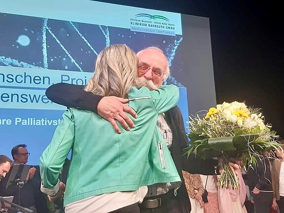 Eine Frau in einer grünen Jacke umarmt einen älteren Mann mit Brille und Bart, der einen bunten Blumenstrauß in der Hand hält. Im Hintergrund sind weitere Personen und ein Banner sichtbar, das Informationen über ein Projekt zu Palliativversorgung zeigt.