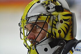 Ein Eishockey-Torwart trägt einen auffälligen Helm mit einem Tigermuster in Gelb und Schwarz. Sein Gesichtsausdruck ist konzentriert und entschlossen, während er auf das Spiel vorbereitet ist. Der Hintergrund ist unscharf, um den Fokus auf den Torwart zu legen.