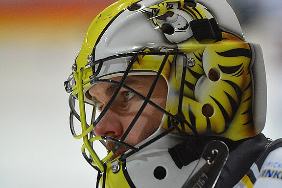 Ein Eishockey-Torwart trägt einen auffälligen Helm mit einem Tigermuster in Gelb und Schwarz. Sein Gesichtsausdruck ist konzentriert und entschlossen, während er auf das Spiel vorbereitet ist. Der Hintergrund ist unscharf, um den Fokus auf den Torwart zu legen.