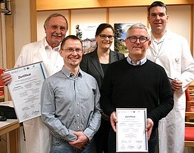 Fünf Personen stehen in einem Raum zusammen. Sie halten Zertifikate in den Händen. Die Gruppe besteht aus vier Männern und einer Frau, alle in formeller Kleidung. Im Hintergrund sind Holzregale und eine Landschaftsabweichung sichtbar.