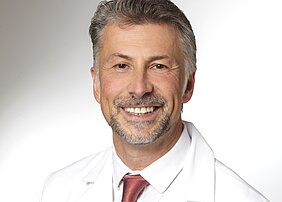 Ein lächelnder Mann trägt einen weißen Arztkittel und eine rote Krawatte. Sein Gesicht zeigt ein freundliches Auftreten. Der Hintergrund ist neutral gehalten, um den Fokus auf den Mann zu legen.