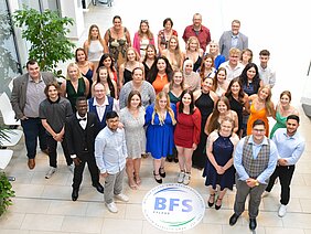 Gruppenfoto von 50 Personen, die in festlicher Kleidung zusammenstehen. Die Gruppe besteht aus Männern und Frauen unterschiedlichen Alters, auf einem hellen Hintergrund mit Pflanzen. Im Vordergrund ist ein Logo mit der Aufschrift "BFS Pflege" zu sehen.
