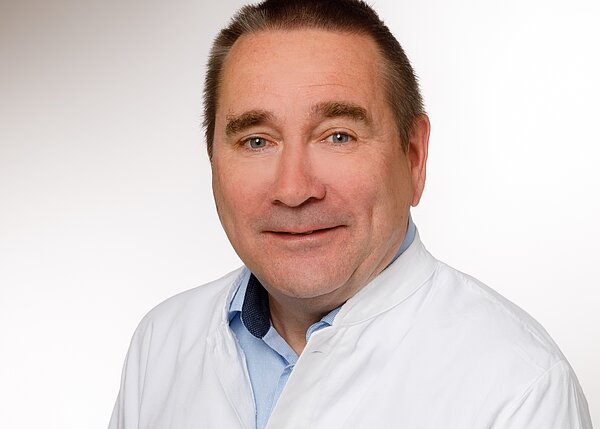 Ein mittelalterlicher Mann mit einem freundlichen Lächeln trägt einen weißen Kittel über einem blauen Hemd. Er schaut in die Kamera und hat kurzes, leicht graues Haar. Der Hintergrund ist hell und unauffällig.