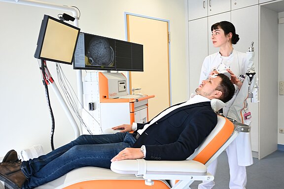 Ein Arzt steht hinter einem Patienten, der auf einer Liege sitzt. Der Patient sieht nervös aus, während der Arzt ein Gerät an seinem Kopf hält. Auf einem Bildschirm sind medizinische Informationen zu erkennen, möglicherweise eine Gehirnaufnahme. Die Umgebung wirkt klinisch und professionell.