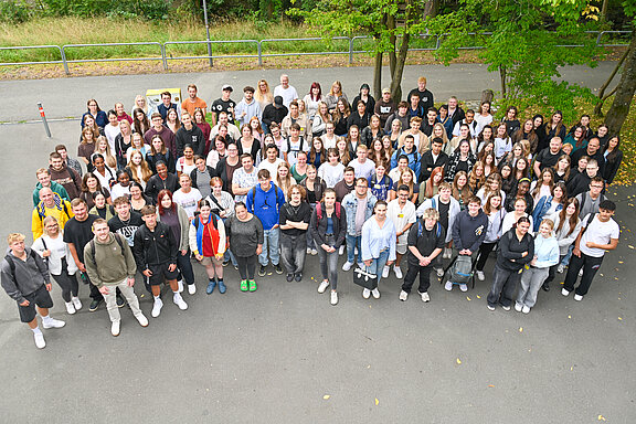 Eine große Gruppe von Menschen steht im Freien, auf einem gepflasterten Platz. Die Personen sind vielfältig gekleidet und lächeln in die Kamera. Bäume und Wiese sind im Hintergrund sichtbar. Die Anordnung der Gruppe vermittelt ein Gefühl von Gemeinschaft und Zusammengehörigkeit.