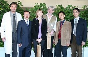 Sechs Personen stehen in einer Gruppe, umgeben von Pflanzen. Sie tragen formelle Kleidung und zeigen verschiedene Ausdrücke. In der Mitte hält eine Frau eine Tasche. Die Szene vermittelt eine Atmosphäre von Zusammenarbeit oder Feierlichkeit.