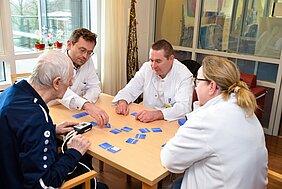 Vier Personen sitzen an einem Tisch und spielen mit blauen Karten. Eine ältere Person, die ein medizinisches Gerät in der Hand hält, wird von drei Fachkräften in weißen Kitteln betreut. Die Atmosphäre wirkt freundlich und kommunikativ, während sie gemeinsam an der Aktivität arbeiten.