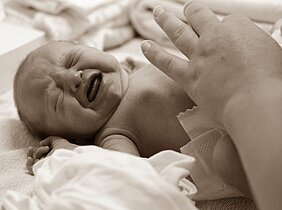 Ein weinendes Baby liegt auf einer weichen Unterlage. Eine Hand berührt sanft das Baby, das Unbehagen zeigt. Die Szene vermittelt eine emotionale Verbindung zwischen dem Baby und der beruhigenden Hand.