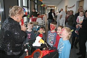 Eine ältere Frau mit glitzerndem Oberteil unterhält eine Gruppe von Kindern, die mit Gesichtern bemalt sind. Die Kinder schauen gespannt zu, während die Frau einen Zauberhut und Requisiten präsentiert. Im Hintergrund sind weitere Personen und festliche Dekorationen zu sehen.