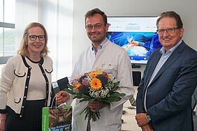 Die Abbildung zeigt drei Personen in einem Raum. Ein Arzt in einem weißen Kittel hält einen Blumenstrauß. Eine Frau lächelt und gibt ihm eine Tragetasche. Der Mann rechts trägt einen Anzug und schaut freundlich. Im Hintergrund ist ein Bildschirm mit medizinischen Bildern sichtbar.