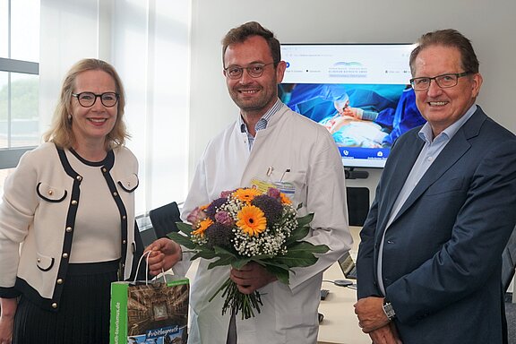 Die Abbildung zeigt drei Personen in einem Raum. Ein Arzt in einem weißen Kittel hält einen Blumenstrauß. Eine Frau lächelt und gibt ihm eine Tragetasche. Der Mann rechts trägt einen Anzug und schaut freundlich. Im Hintergrund ist ein Bildschirm mit medizinischen Bildern sichtbar.