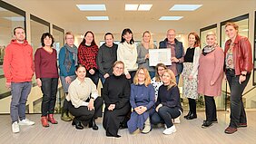 Eine Gruppe von 15 Personen steht und sitzt in einem modernen Büro. Einige Personen halten eine Urkunde. Die Gruppe zeigt Freude und Zusammenhalt. Die Umgebung ist hell mit großen Fenstern und einen offenen Raum.