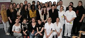 Eine Gruppe von etwa 30 Personen steht in einem Raum. Die Mehrheit trägt alltägliche Kleidung, einige tragen weiße Arbeitskleidung. Im Hintergrund sind Banner und eine Präsentation sichtbar. Die Stimmung wirkt freundlich und gemeinschaftlich.