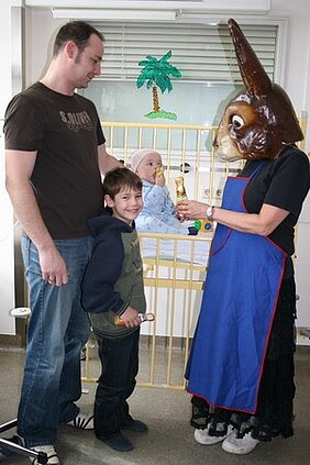 Ein Mann und ein Junge stehen neben einem Bettchen, in dem ein Baby liegt. Eine Person in einer ungewöhnlichen Maske reicht dem Jungen eine kleine Statue. Der Hintergrund zeigt einen einfachen Raum mit einem Fenster und einer Palme als Dekoration.