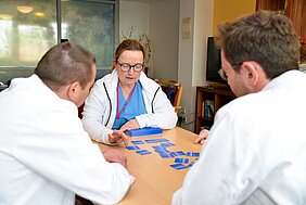 Drei Personen sitzen an einem Tisch und spielen mit blauen Karten. Eine Frau in blauer Kleidung erklärt das Spiel, während die anderen beiden aufmerksam zuhören und ihre Hände auf dem Tisch haben. Der Raum wirkt freundlich und einladend.