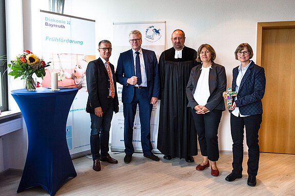 Eine Gruppe von fünf Personen steht in einem Raum. Zwei Männer tragen Anzüge, einer ist in Robe gekleidet. Die Frauen stehen in eleganter Kleidung. Im Hintergrund sind Banner der Diakonie Bayreuth und einer Förderorganisation sichtbar. Auf einem Tisch stehen Blumen und Kerzen.