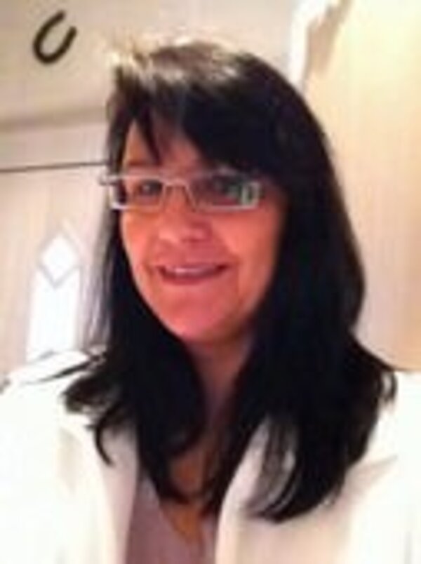 Eine lächelnde Person mit langen, schwarzen Haaren trägt eine Brille und einen weißen Blazer. Der Hintergrund zeigt eine Tür mit Fensterelementen. Die Person vermittelt ein freundliches und professionelles Erscheinungsbild.