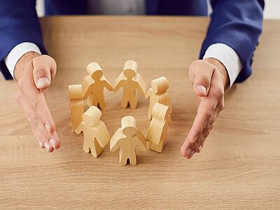 Ein paar Holzfiguren stehen händehaltend in einem Kreis auf einem Tisch. Die Hände eines Mannes, der einen Anzug trägt, umrahmen die Figuren schützend, was Gemeinschaft und Schutz symbolisiert. Menschenrechte.jpg