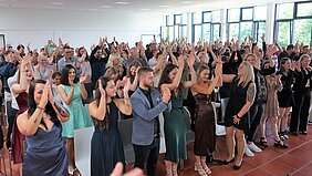 Eine große Gruppe von Menschen steht in einem Raum und applaudiert begeistert. Die Anwesenden tragen verschiedene formelle Kleidung und zeigen Freude. Die Atmosphäre ist festlich und es scheint ein besonderes Ereignis zu feiern. Tageslicht strömt durch große Fenster.