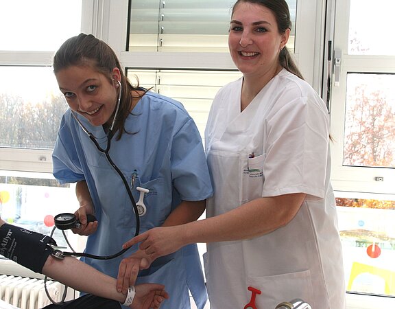 Zwei Pflegekräfte stehen in einem hellen Raum. Eine Krankenschwester mit einem Stethoskop hört den Puls eines Patienten, während die andere lächelnd einen Arm des Patienten mit einer Manschette für die Blutdruckmessung hält.
