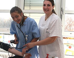 Zwei Pflegekräfte stehen in einem hellen Raum. Eine Krankenschwester mit einem Stethoskop hört den Puls eines Patienten, während die andere lächelnd einen Arm des Patienten mit einer Manschette für die Blutdruckmessung hält.