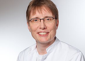 Ein lächelnder Arzt oder eine Ärztin mit kurzen, braunen Haaren trägt eine weiße medizinische Jacke und Brille. Der Hintergrund ist hell und neutral. Die Person vermittelt einen freundlichen und professionellen Eindruck.