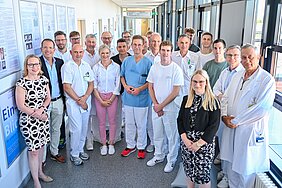 Eine Gruppe von medizinischem Personal steht in einem Flur. Alle tragen Arbeitskleidung, darunter weiße Kittel. Im Hintergrund sind Informationsplakate und Fenster sichtbar. Die Stimmung wirkt freundlich und professionell.