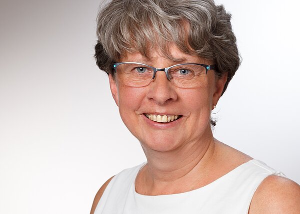 Porträt einer lächelnden Frau mit kurzen, grauen Haaren, die eine brille trägt. Sie hat einen freundlichen Ausdruck und trägt ein ärmelloses, helles Oberteil. Der Hintergrund ist neutral und hell.