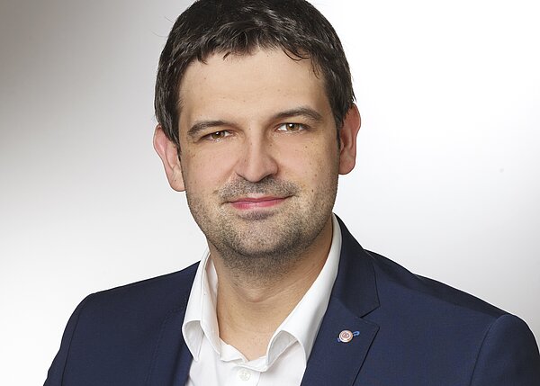 Ein Porträt eines Mannes mit kurzen, dunklen Haaren, der in einem dunkelblauen Blazer und einem weißen Hemd gekleidet ist. Er lächelt leicht und blickt direkt in die Kamera. Der Hintergrund ist hell und einfarbig.