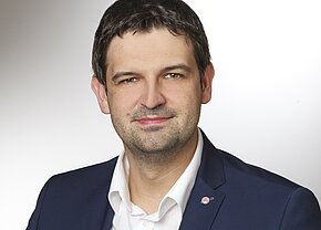 Ein Porträt eines Mannes mit kurzen, dunklen Haaren, der in einem dunkelblauen Blazer und einem weißen Hemd gekleidet ist. Er lächelt leicht und blickt direkt in die Kamera. Der Hintergrund ist hell und einfarbig.