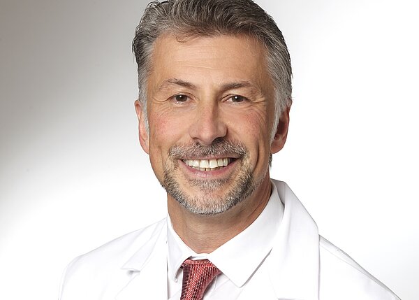 Ein lächelnder Arzt in weißem Kittel mit einem roten Hemd darunter. Sein Haarschnitt ist kurz, und er hat einen gepflegten Bart. Der Hintergrund ist hell und schlicht, was den Fokus auf sein freundliches Gesicht lenkt.