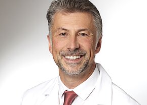 Ein lächelnder Arzt in weißem Kittel mit einem roten Hemd darunter. Sein Haarschnitt ist kurz, und er hat einen gepflegten Bart. Der Hintergrund ist hell und schlicht, was den Fokus auf sein freundliches Gesicht lenkt.