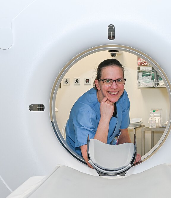 Eine lächelnde Fachkraft in blauer Arbeitskleidung steht am Eingang eines CT-Scanners. Der Raum im Hintergrund zeigt medizinische Geräte und Materialien. Die Person vermittelt eine freundliche und einladende Stimmung.